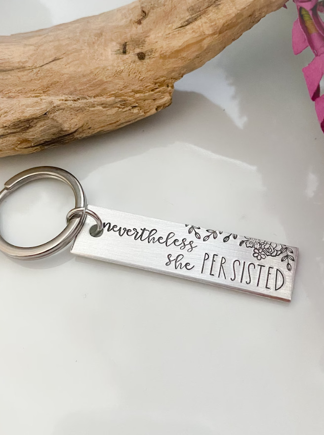 Silver keychain inscribed with saying Nevertheless She Persisted