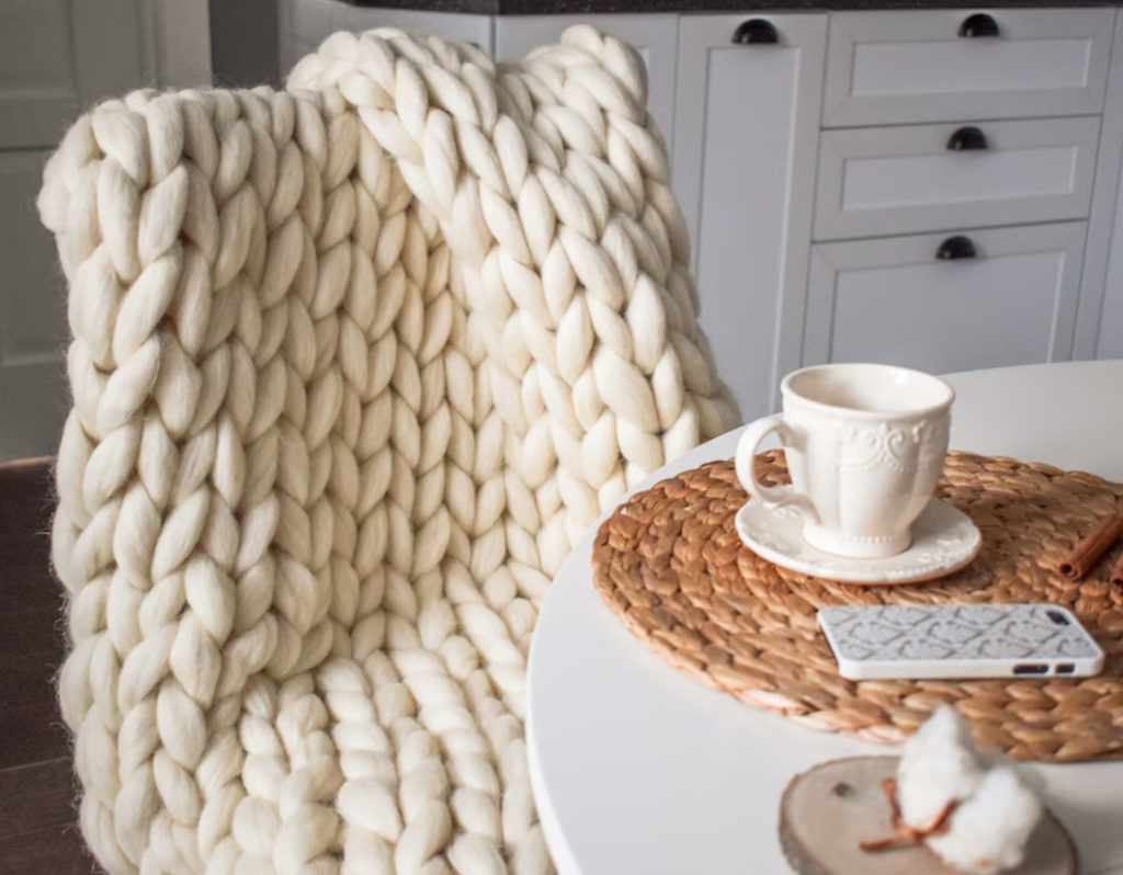 Chunky Knit Merino Wool Blanket draped over chair next to table with coffee cup and cell phone